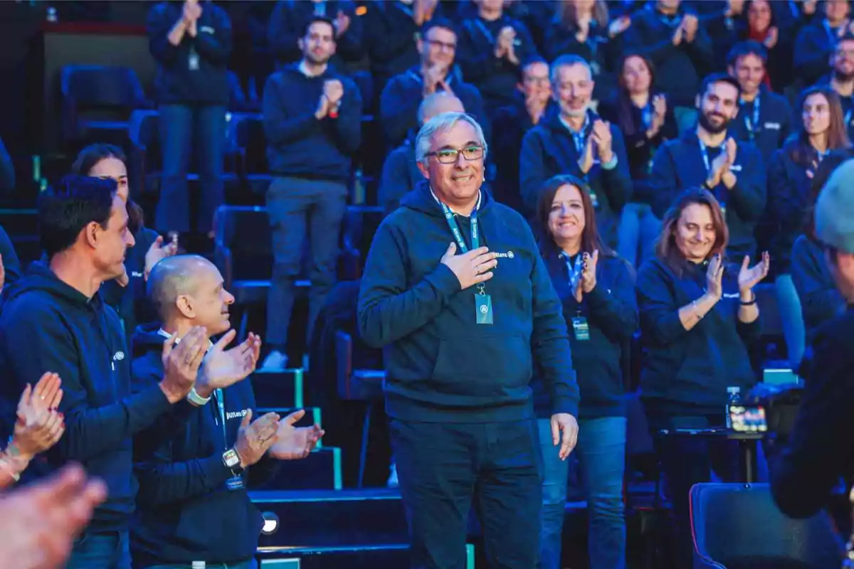 Un hombre de pie en un auditorio rodeado de personas aplaudiendo, todos vistiendo sudaderas azules.