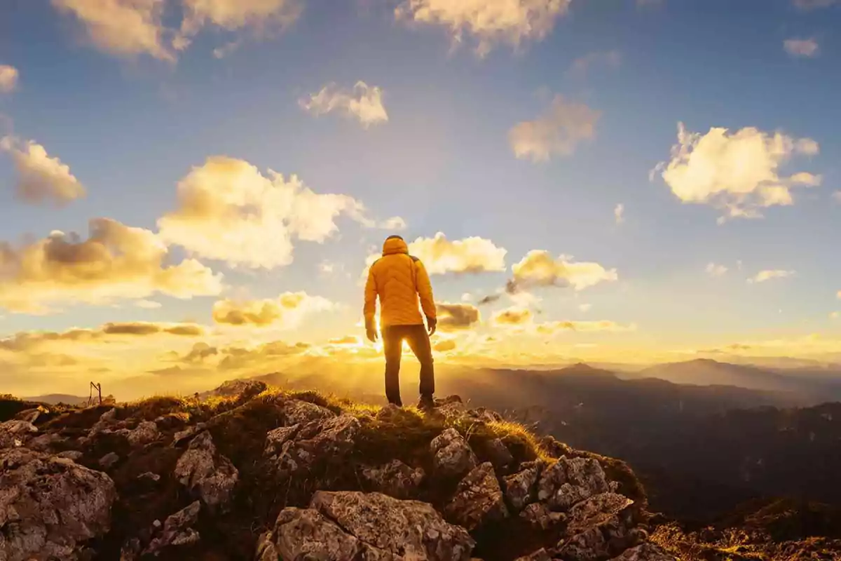 Persona con chaqueta amarilla de pie sobre una montaña rocosa al atardecer con el sol brillando en el horizonte y nubes en el cielo.