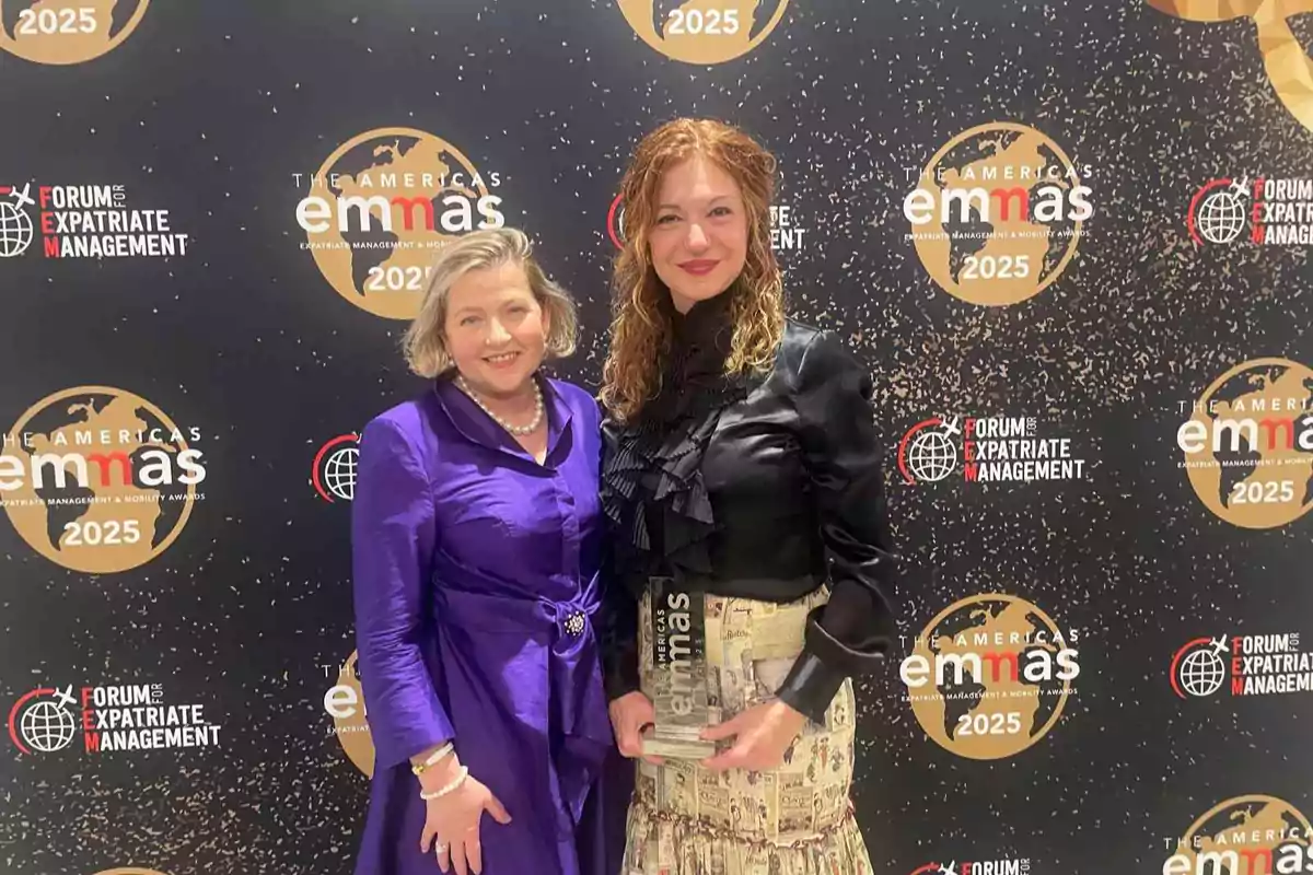 Dos mujeres sonríen frente a un fondo de premiación de los Americas EMMAs 2025, una de ellas sostiene un trofeo