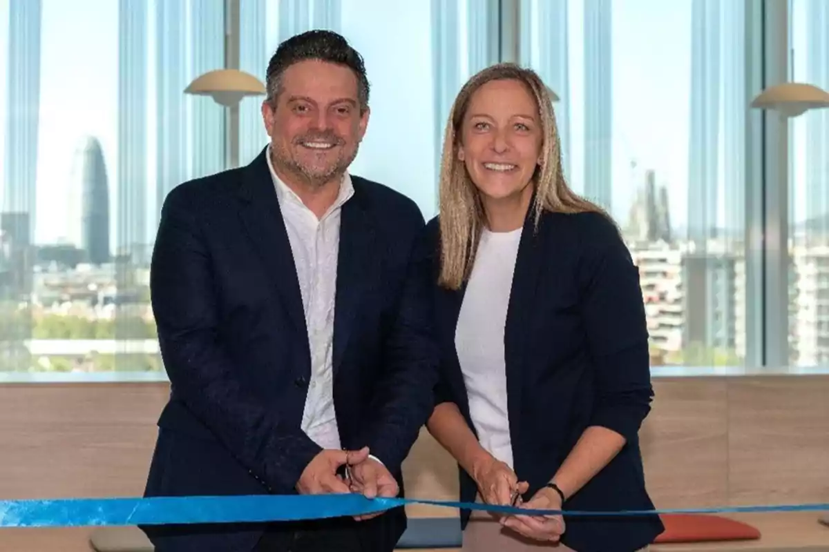 Dos personas sonriendo mientras cortan una cinta azul en una ceremonia de inauguración con un fondo de edificios y ventanas.