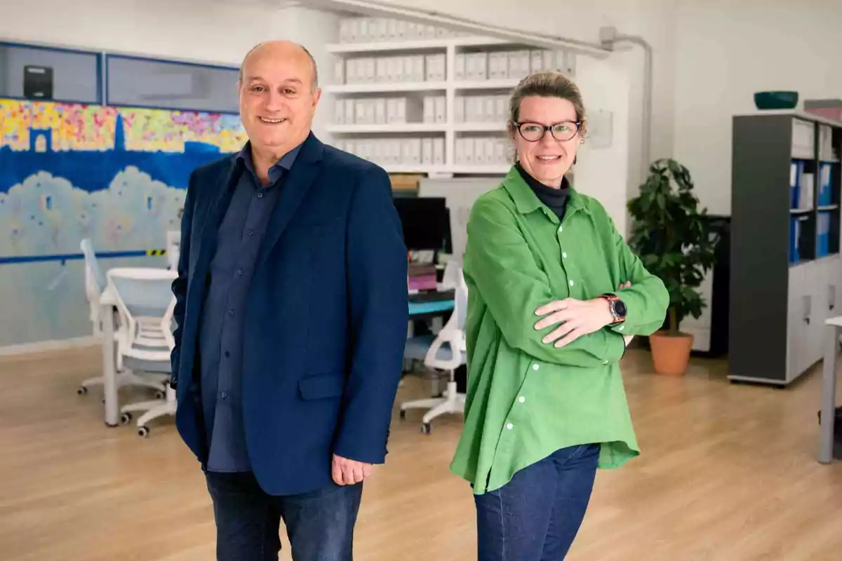 Dos personas posando en una oficina moderna con estanterías y una planta en el fondo.