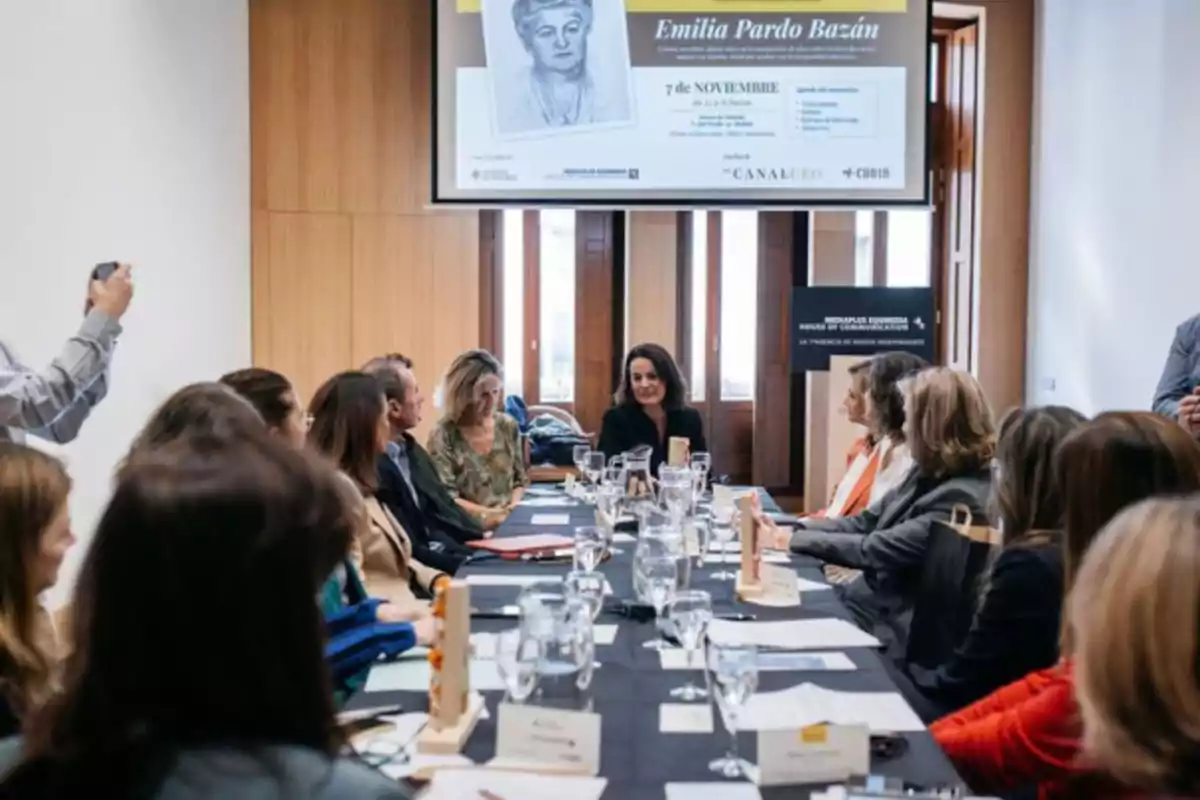 Un grupo de personas está reunido alrededor de una mesa en una sala de conferencias con un cartel de Emilia Pardo Bazán en la pared.