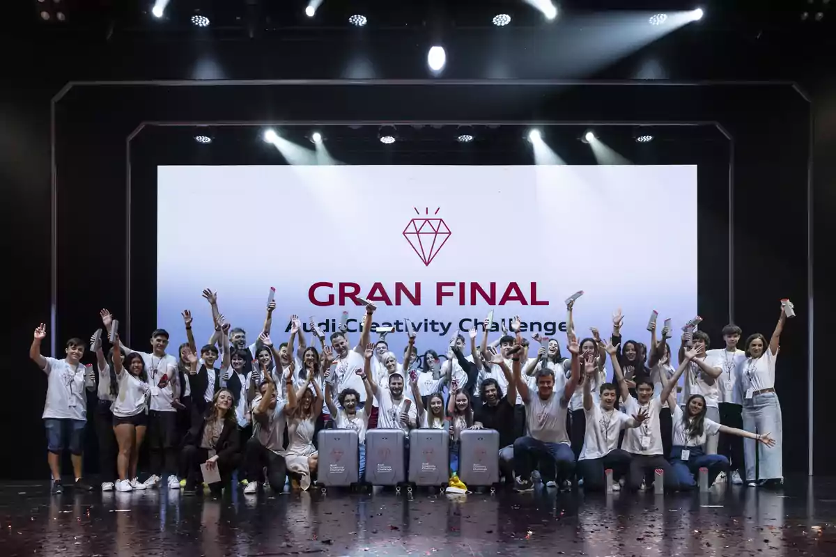 Grupo de jóvenes celebrando en un escenario durante la gran final de un concurso de creatividad, con camisetas blancas y trofeos en la mano, frente a una pantalla que dice Gran Final Audi Creativity Challenge.