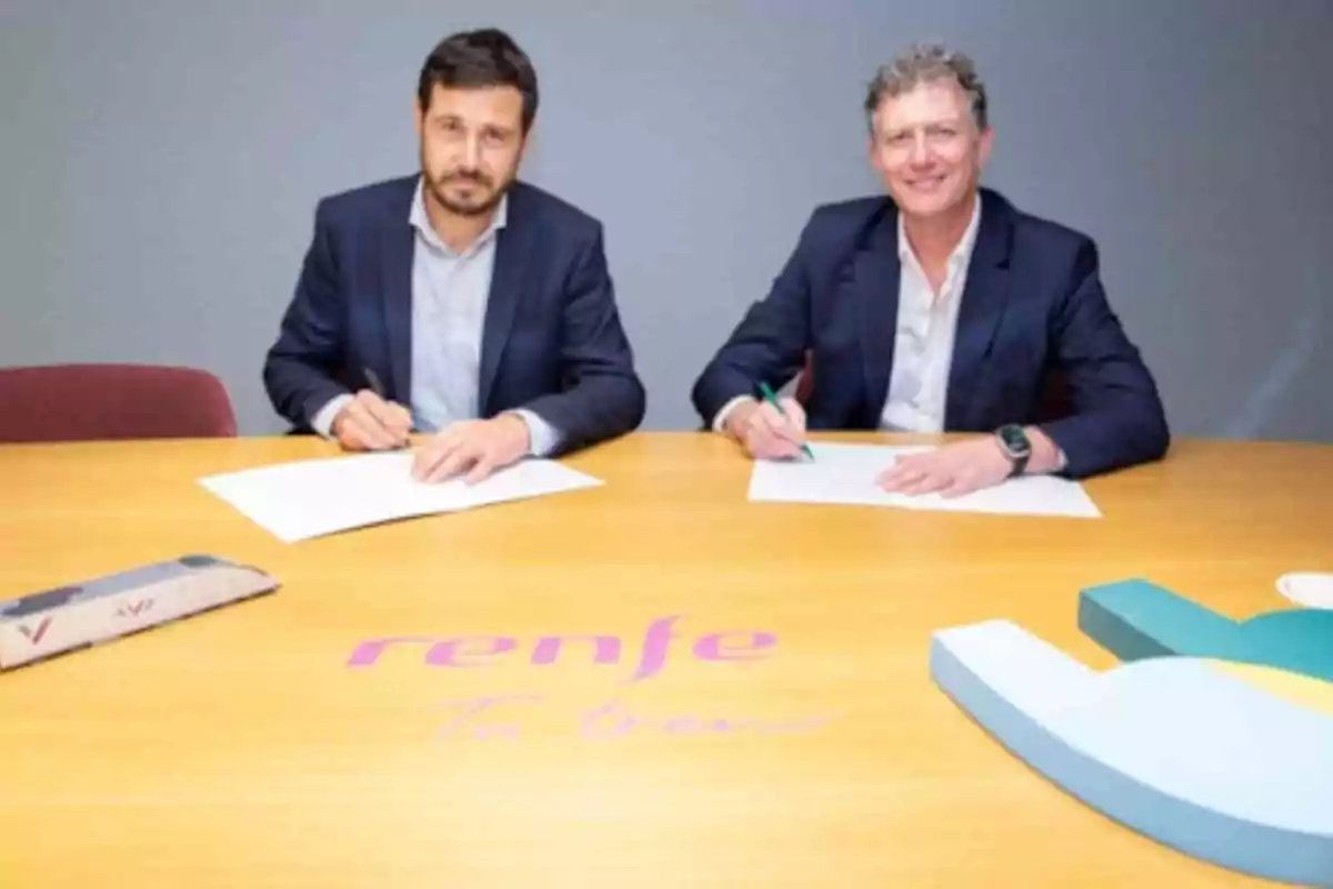 Dos hombres sentados en una mesa firmando documentos con el logo de Renfe visible en la superficie.