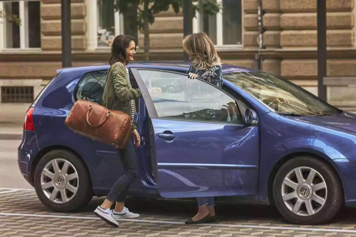 Dos mujeres conversan junto a un auto azul en una calle urbana.