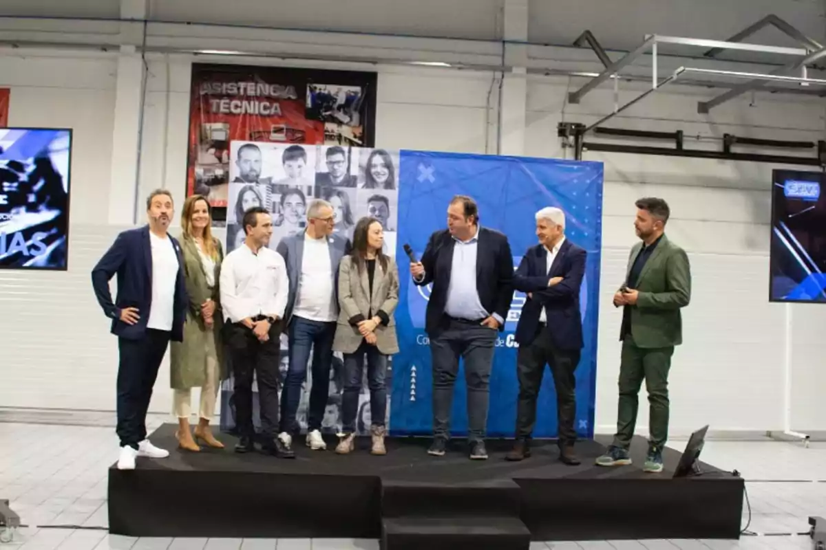 Un grupo de personas está de pie en un escenario interior durante un evento, con pantallas y carteles de fondo.