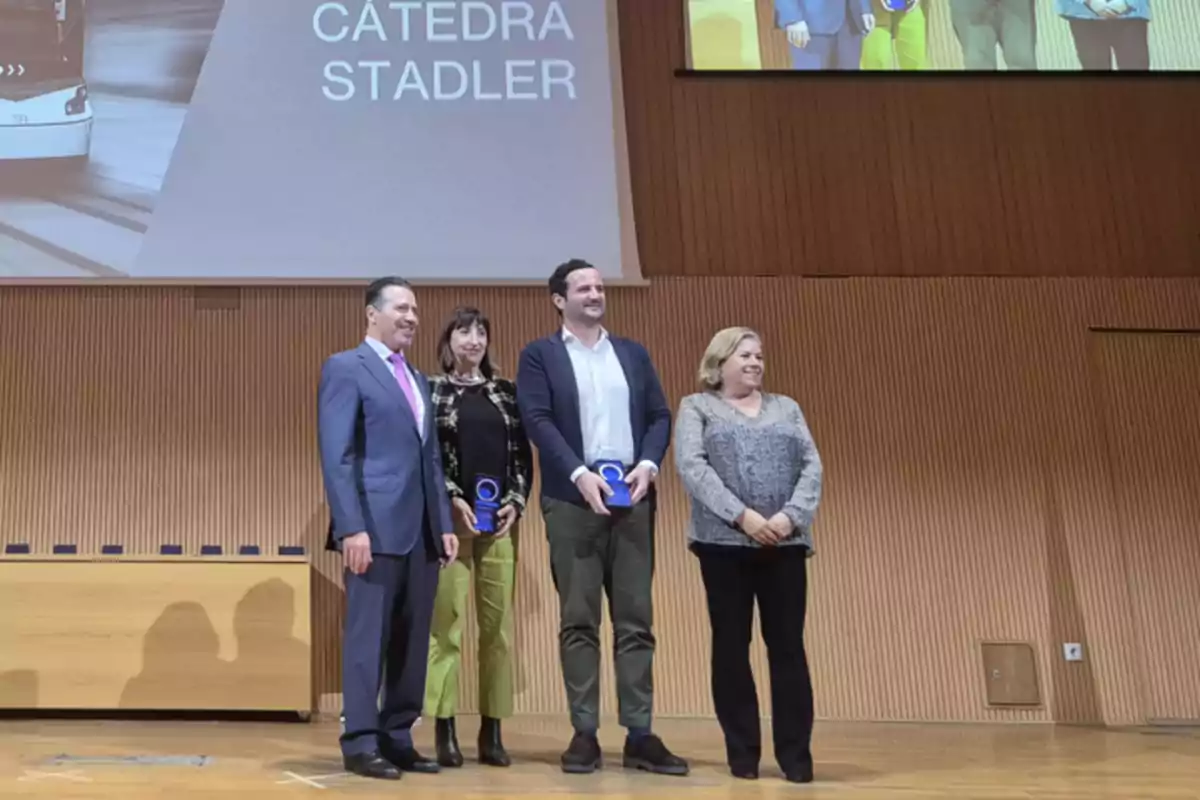 Cuatro personas posan en un escenario durante un evento de la Cátedra Stadler, dos de ellas sostienen trofeos.