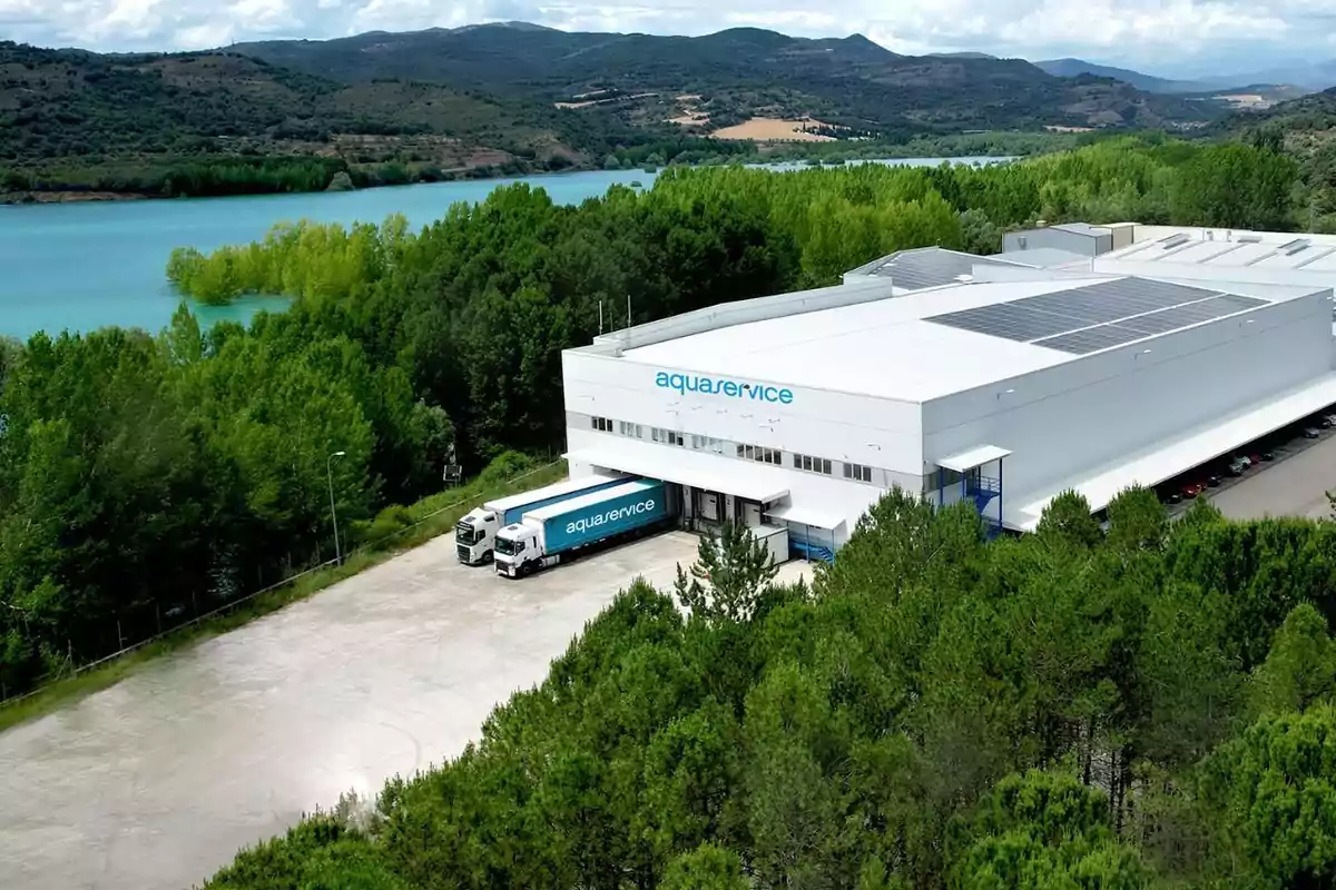 Vista aérea de una nave industrial blanca de Aquaservice rodeada de árboles y cerca de un lago con montañas al fondo