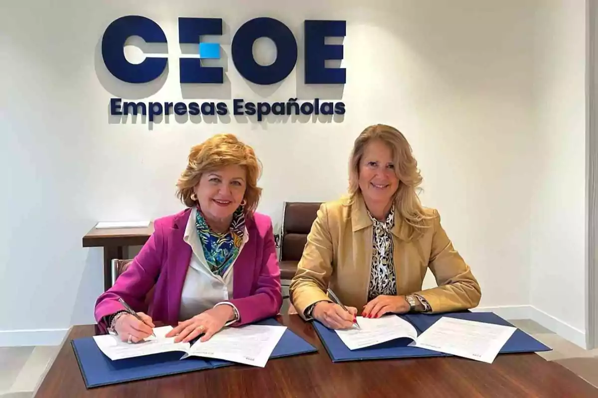 Dos mujeres sonrientes firman documentos en una mesa de oficina con el logo de CEOE Empresas Españolas en la pared de fondo