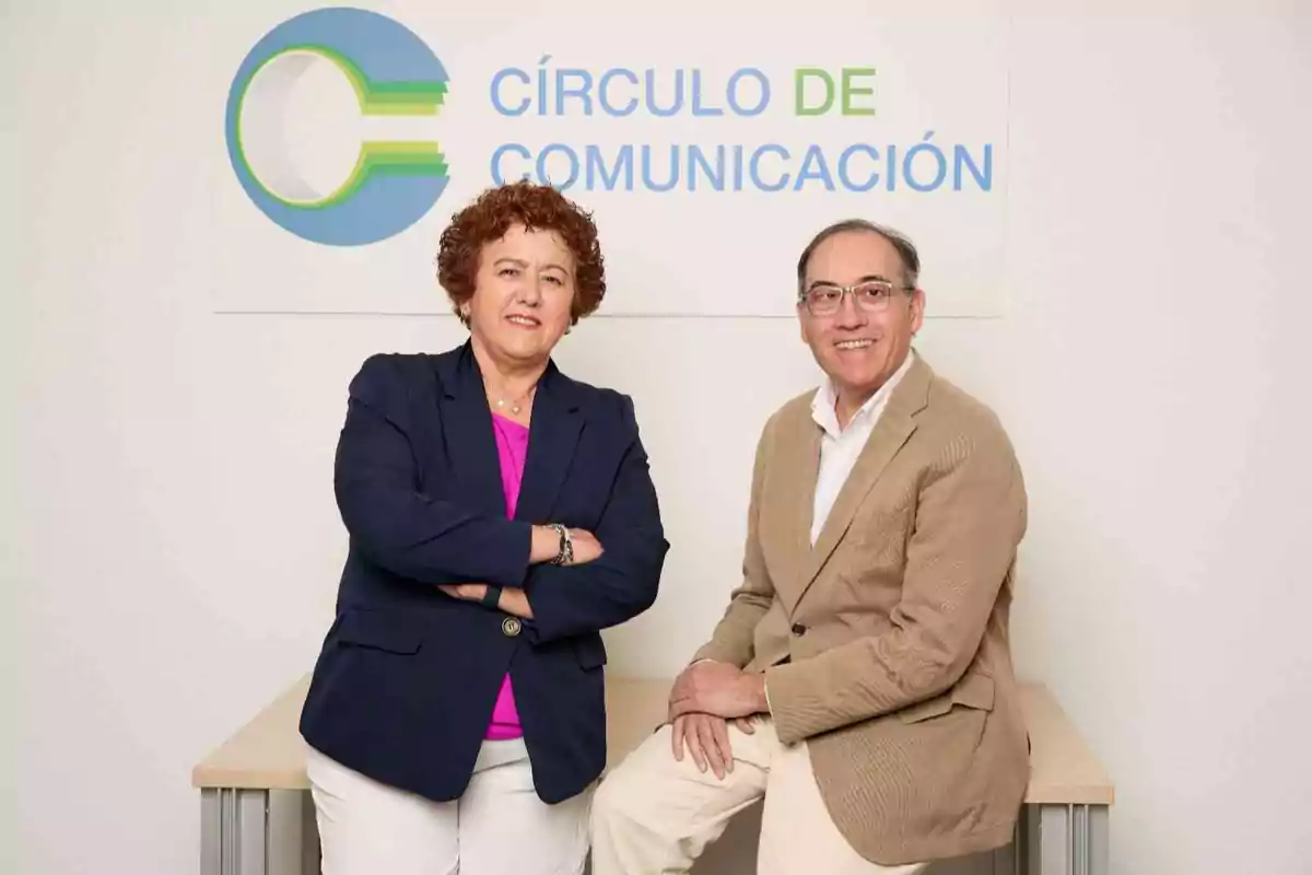 Dos personas posan frente a un cartel que dice Círculo de Comunicación, una de ellas está de pie con los brazos cruzados y la otra está sentada sobre una mesa, ambos sonríen y visten ropa formal.