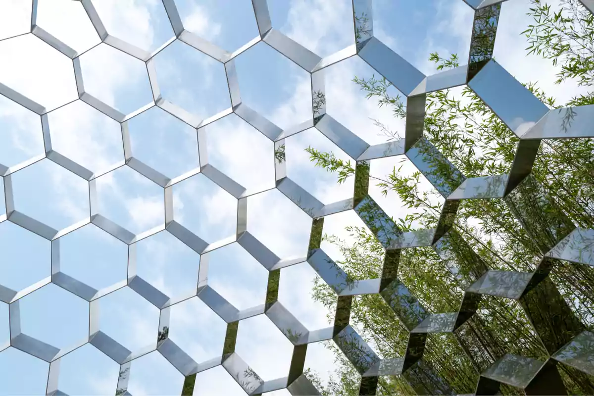 Estructura metálica con forma de panal reflejando el cielo azul y plantas verdes