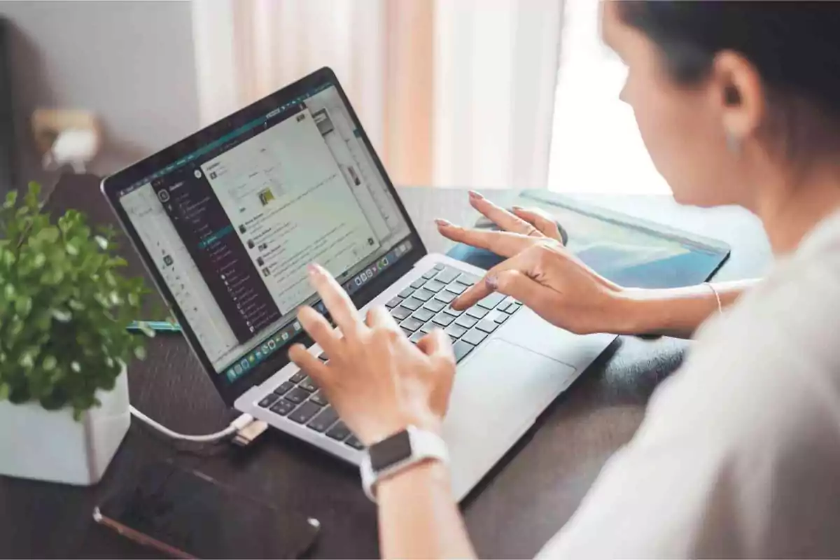 Persona trabajando en una computadora portátil en un escritorio con una planta y un teléfono móvil al lado.