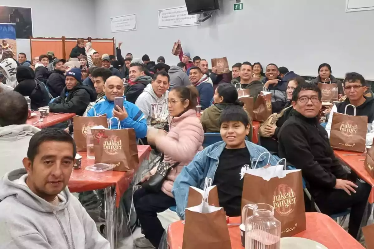 Un grupo de personas se encuentra reunido en un comedor, muchas de ellas sosteniendo bolsas de papel con el logo de "naked burger".