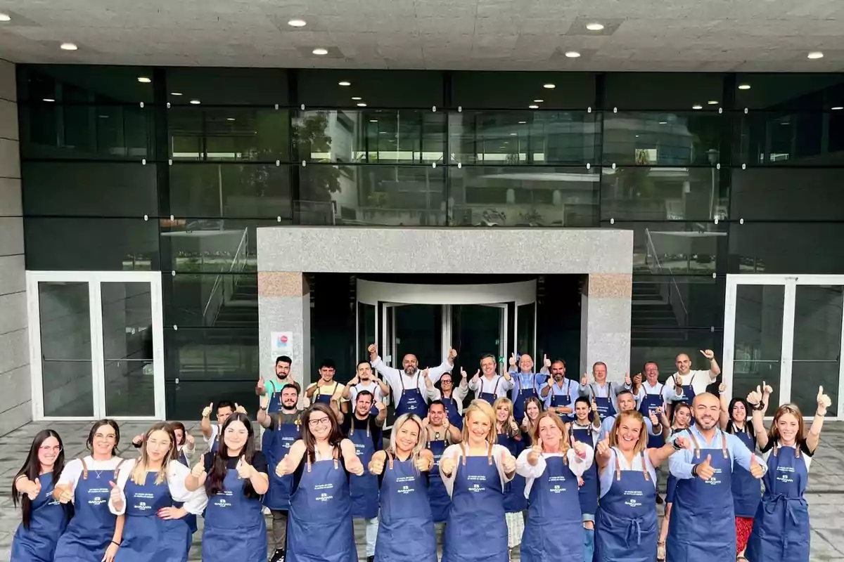 Un grupo de personas con delantales azules posando frente a un edificio moderno con ventanas de vidrio, todas levantando el pulgar en señal de aprobación.