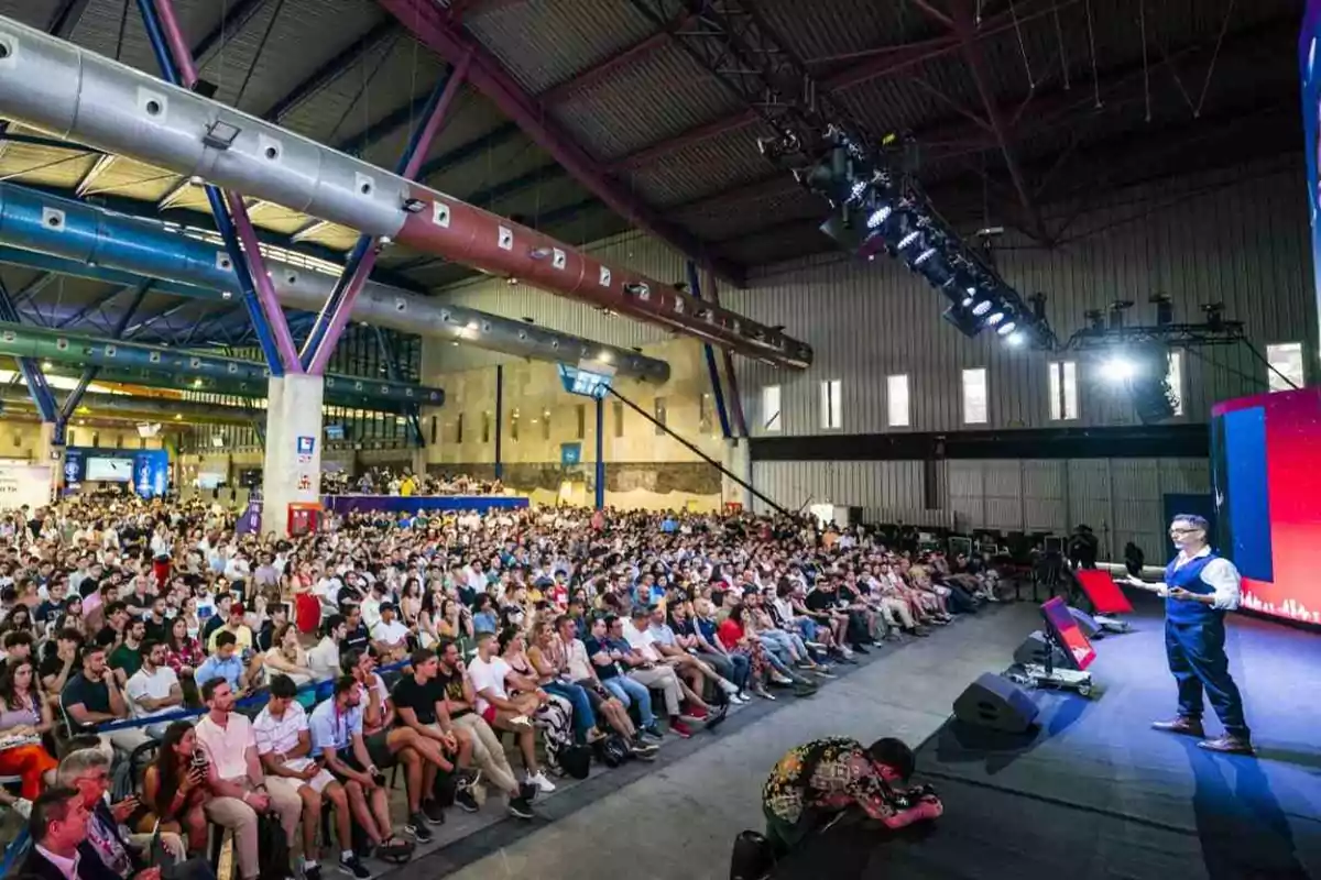 Una multitud de personas asistiendo a una conferencia en un gran auditorio con un orador en el escenario.