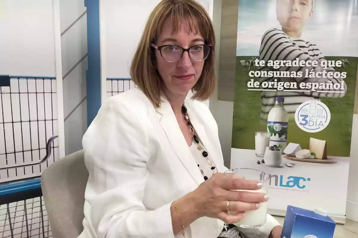 Una mujer con gafas y chaqueta blanca sostiene un vaso de leche frente a un cartel que promueve el consumo de lácteos de origen español.