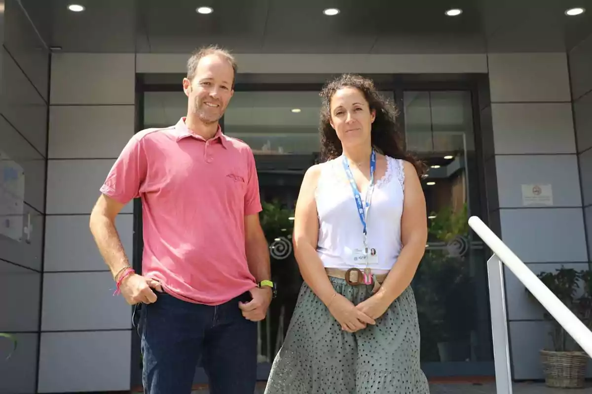 Dos personas posando frente a un edificio moderno con paredes de metal y luces en el techo.