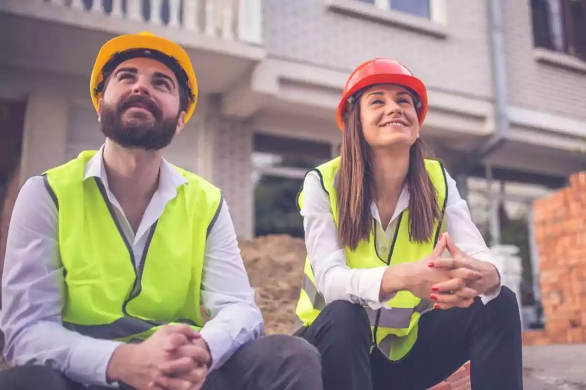 Dos trabajadores de la construcción con chalecos reflectantes y cascos, uno amarillo y otro rojo, sentados y mirando hacia arriba en un sitio de construcción.