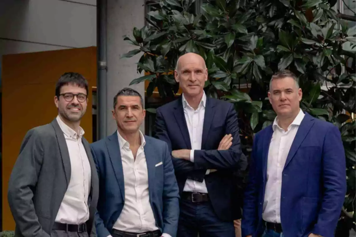 Un grupo de cuatro hombres vestidos con trajes posan juntos frente a un fondo de plantas.