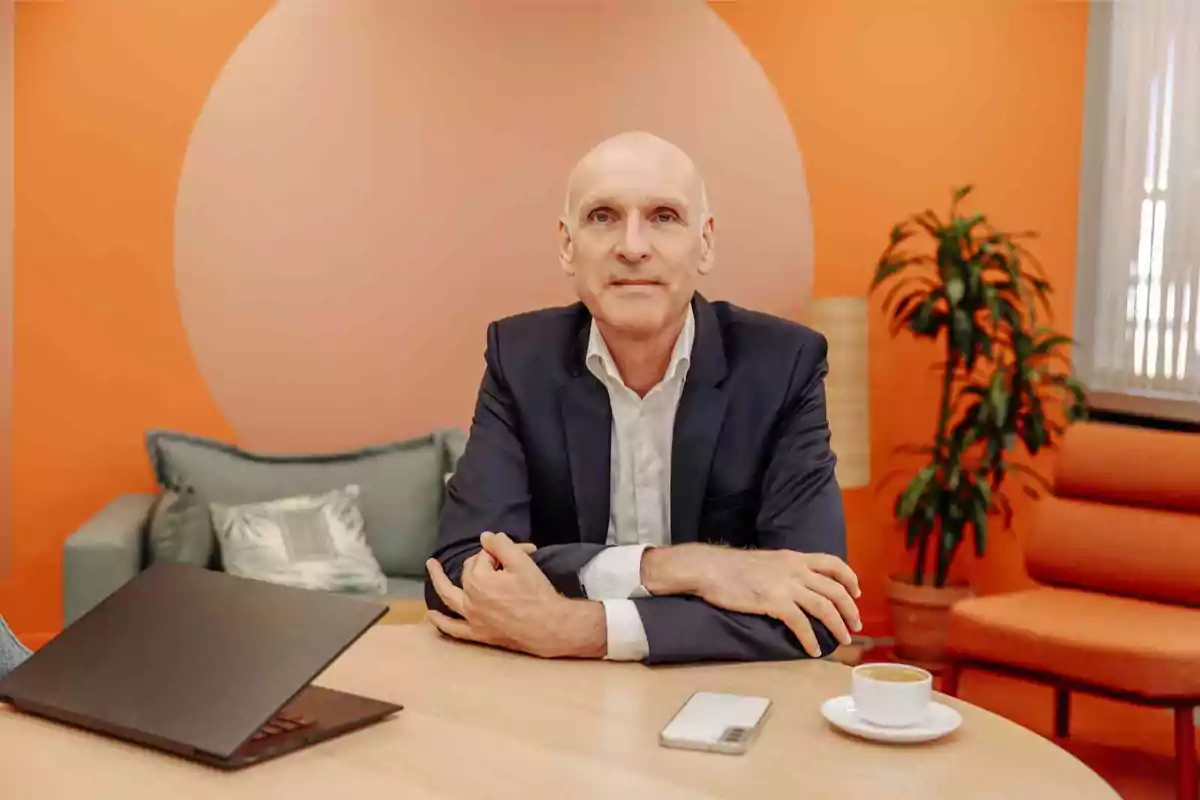 Un hombre calvo con traje oscuro y camisa blanca está sentado en una mesa con un portátil, un teléfono y una taza de café, en un entorno con decoración en tonos naranjas y plantas.