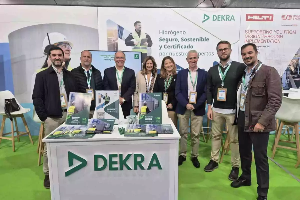 Un grupo de personas posando frente a un stand de DEKRA en un evento, con folletos y materiales promocionales sobre hidrógeno seguro y sostenible.