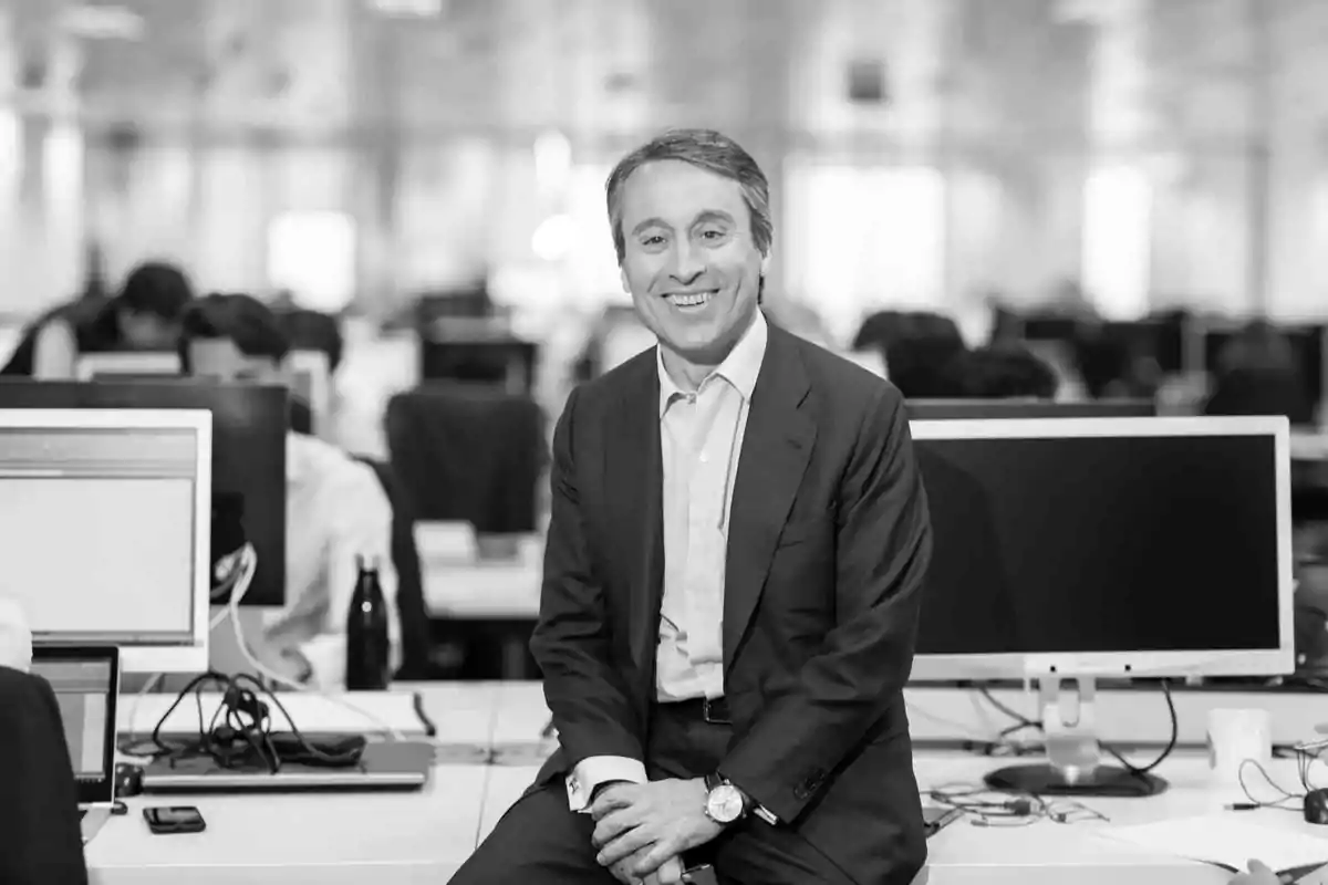 Hombre de traje sonriendo mientras está sentado sobre un escritorio en una oficina moderna con varias computadoras y personas trabajando al fondo