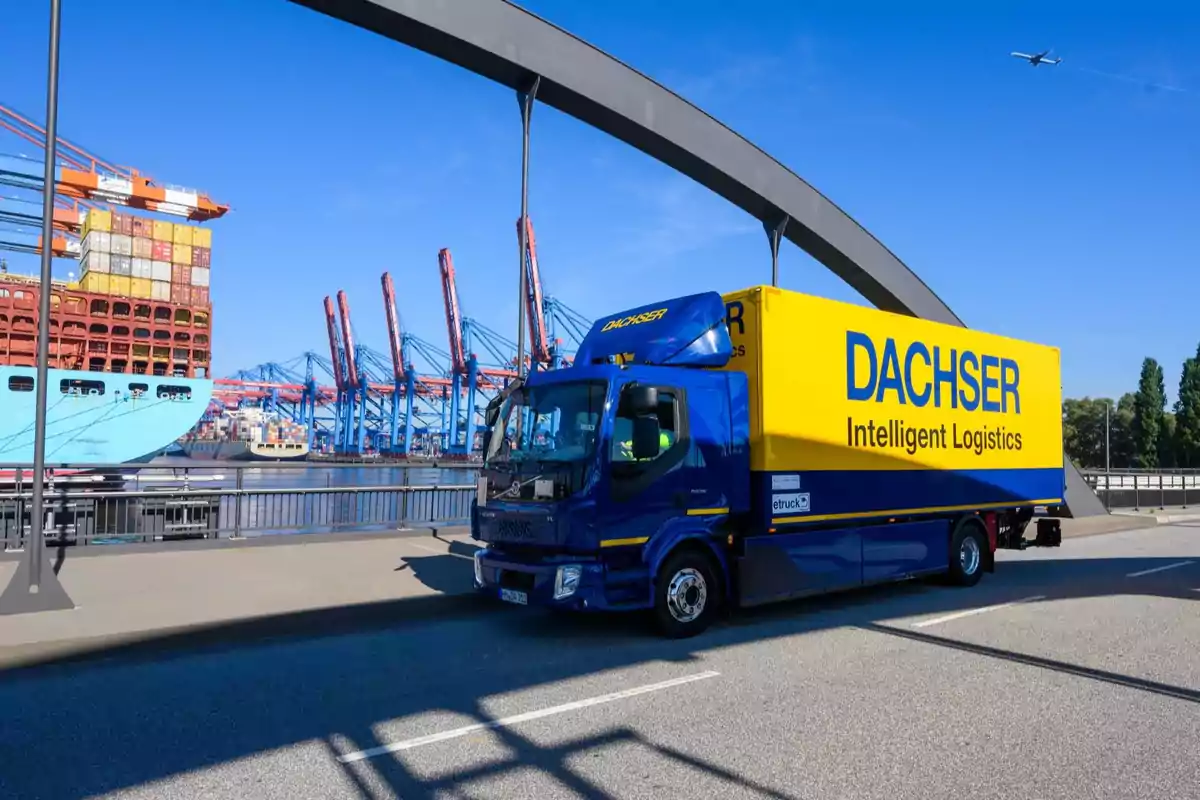 Un camión de logística de color azul y amarillo cruza un puente cerca de un puerto con grúas y un barco de carga.