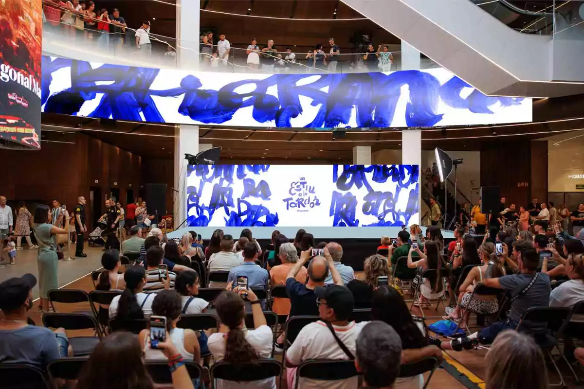 Un evento en un centro comercial con una pantalla grande y muchas personas sentadas observando.