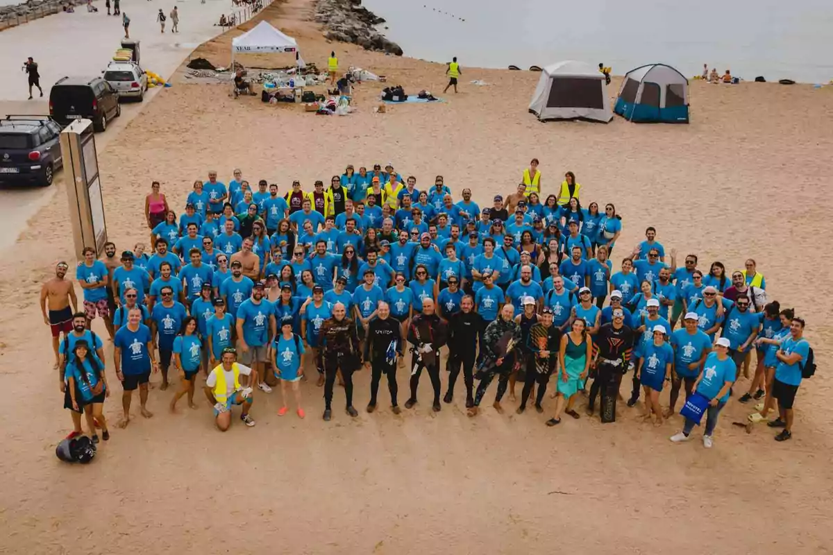 Un grupo grande de personas con camisetas azules se encuentra en una playa, algunas tiendas de campaña y un puesto están en el fondo.