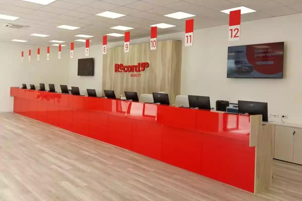 Mostrador de atención al cliente con doce estaciones numeradas y computadoras, con el logo de Record Go Mobility en la pared y una pantalla de televisión.