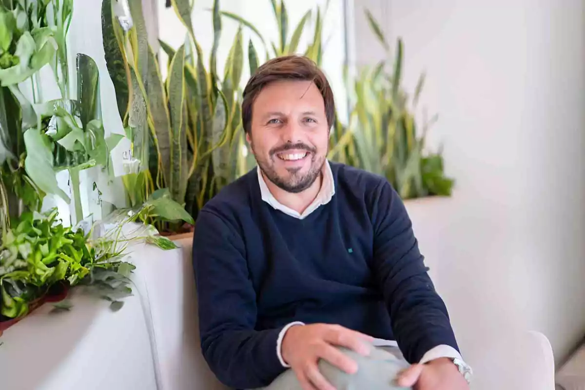 Un hombre sonriente sentado en un sofá rodeado de plantas verdes.