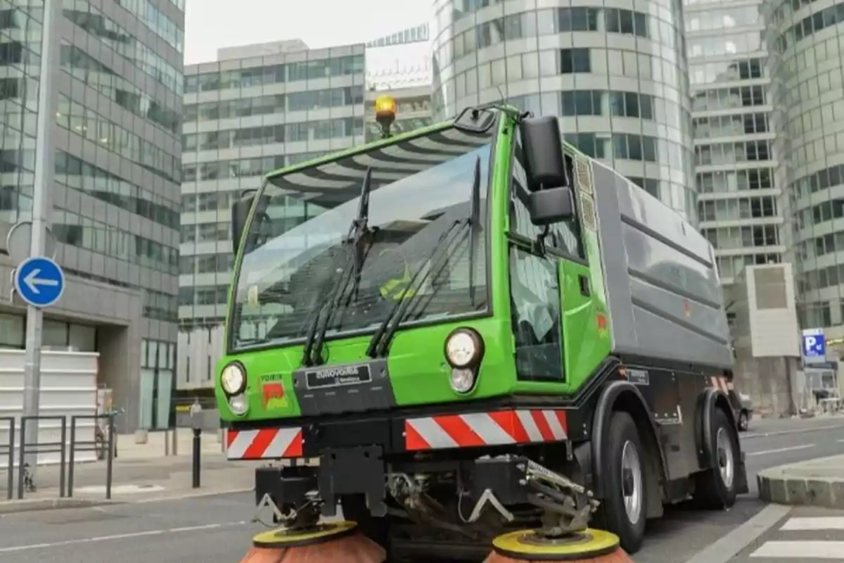 Un camión de limpieza verde en una calle de una ciudad con edificios altos de fondo.