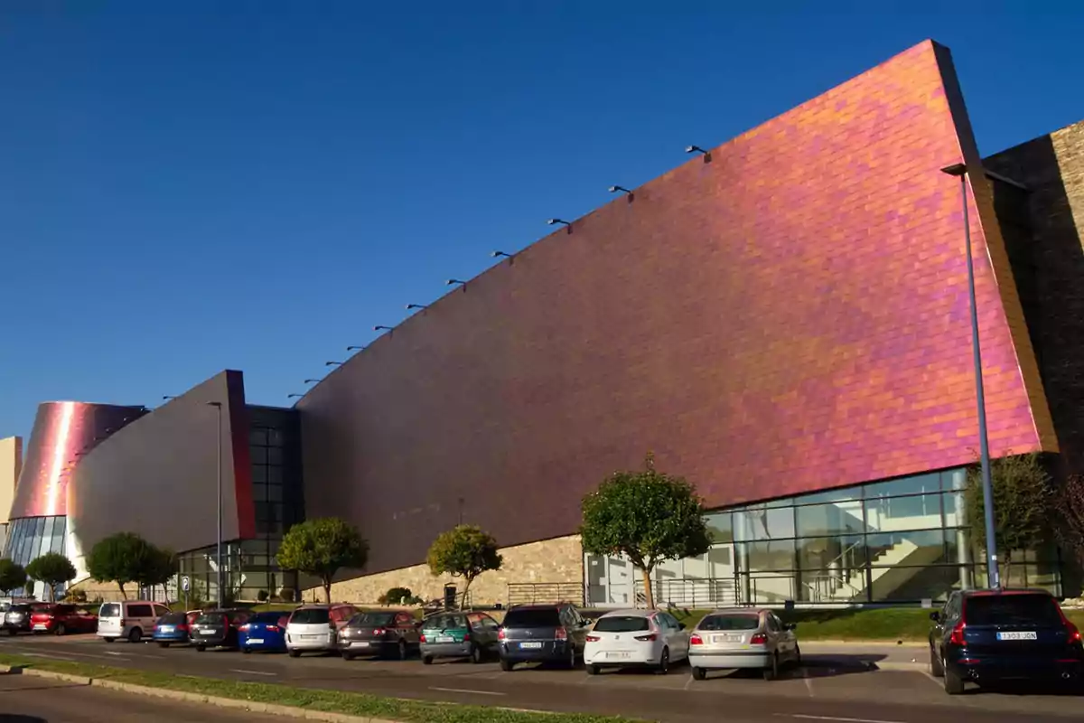 Edificio moderno con fachada de colores iridiscentes y estacionamiento lleno de autos en primer plano.