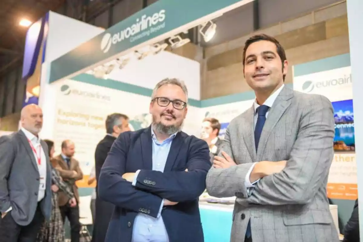 Dos hombres de pie con los brazos cruzados en un stand de una feria comercial de Euroairlines, con varias personas y carteles en el fondo.