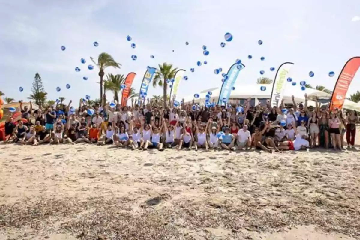 Un grupo grande de personas está reunido en la playa, muchas de ellas levantando los brazos y lanzando pelotas azules al aire, con varias banderas de colores en el fondo y palmeras alrededor.