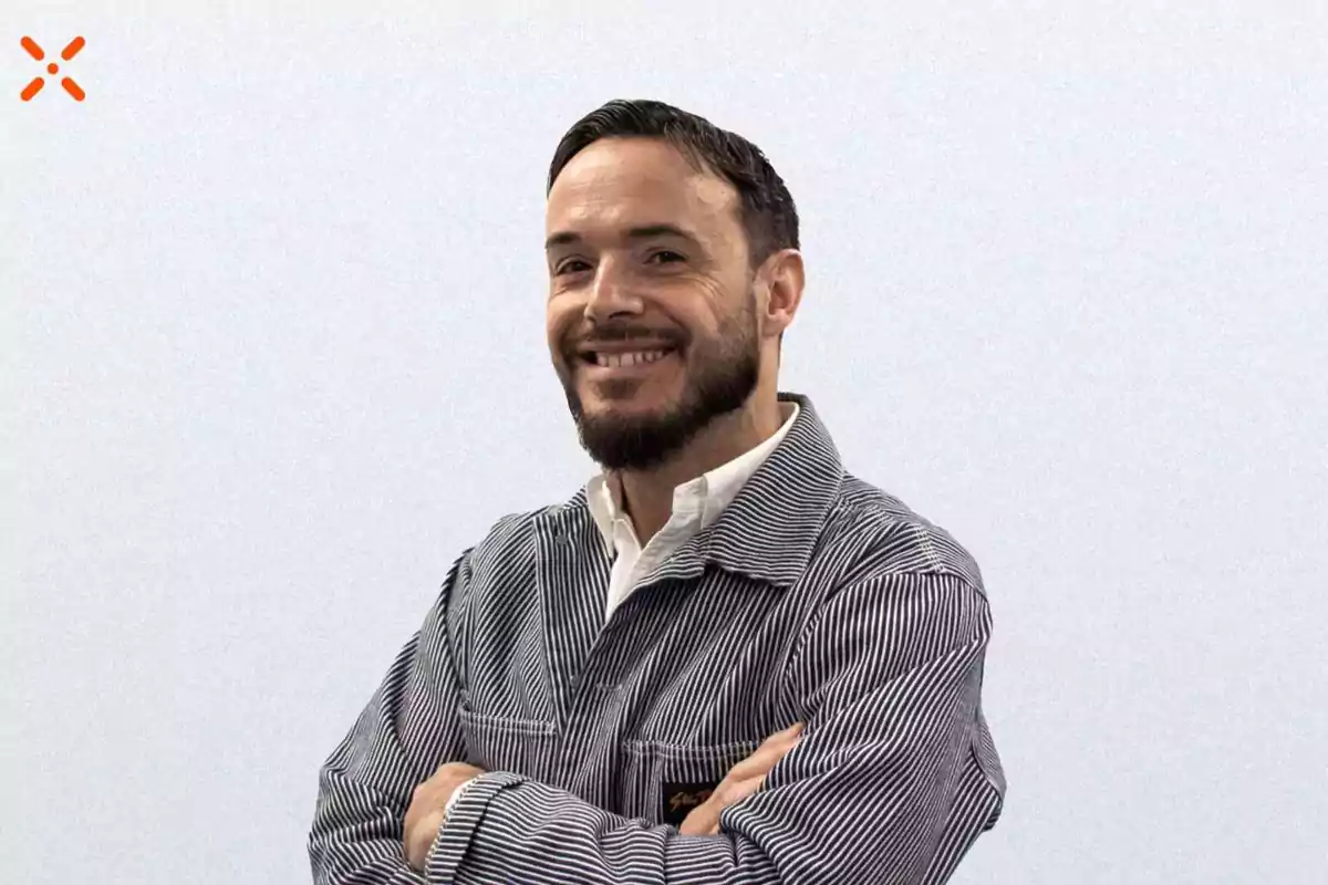 Un hombre con barba y cabello corto sonríe mientras cruza los brazos, vistiendo una chaqueta de rayas sobre una camisa blanca, con un fondo claro.