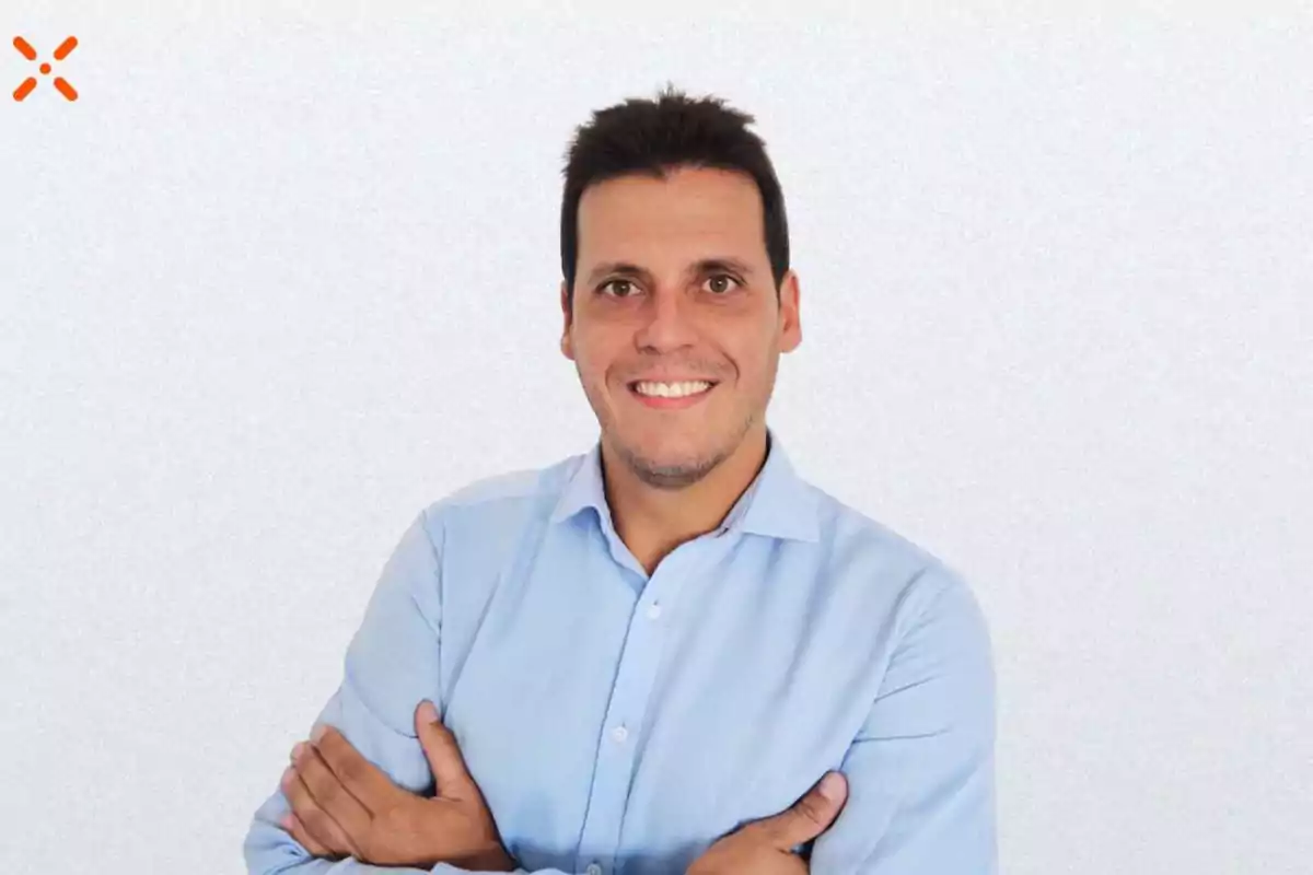 Hombre con camisa azul claro sonriendo y con los brazos cruzados frente a un fondo blanco.