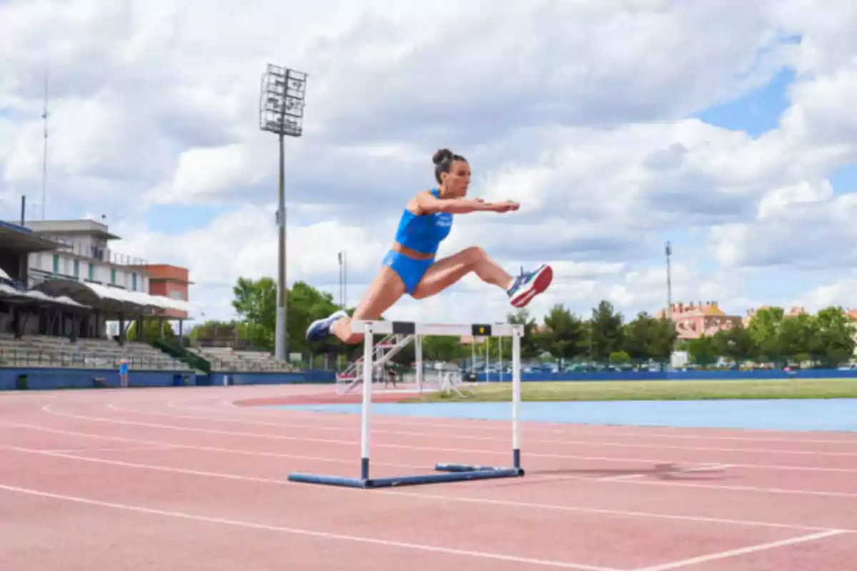 Una atleta en uniforme azul está saltando una valla en una pista de atletismo al aire libre bajo un cielo parcialmente nublado.