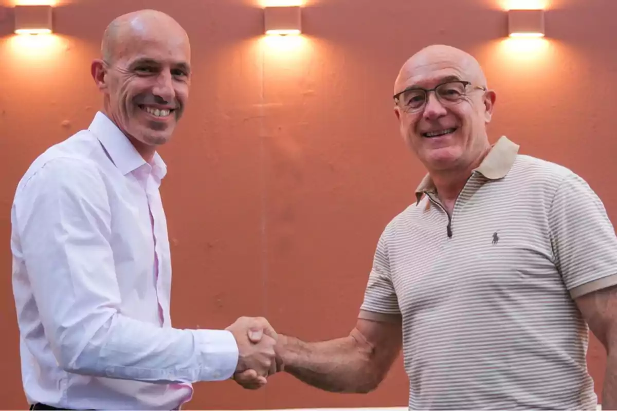Dos hombres sonrientes se dan la mano frente a una pared color salmón con luces encendidas en la parte superior.