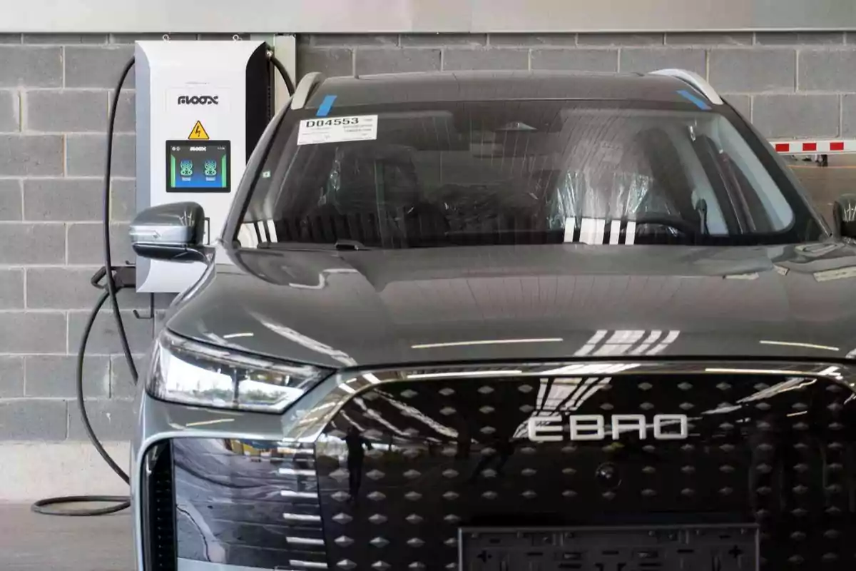 Automóvil eléctrico de color gris estacionado frente a una estación de carga en un entorno industrial