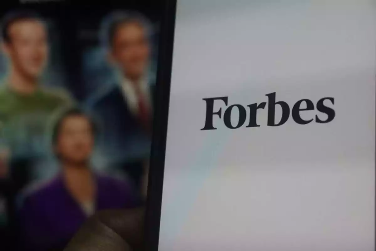 Una pantalla de un dispositivo móvil muestra el logotipo de Forbes, mientras que en el fondo se ven imágenes borrosas de personas.