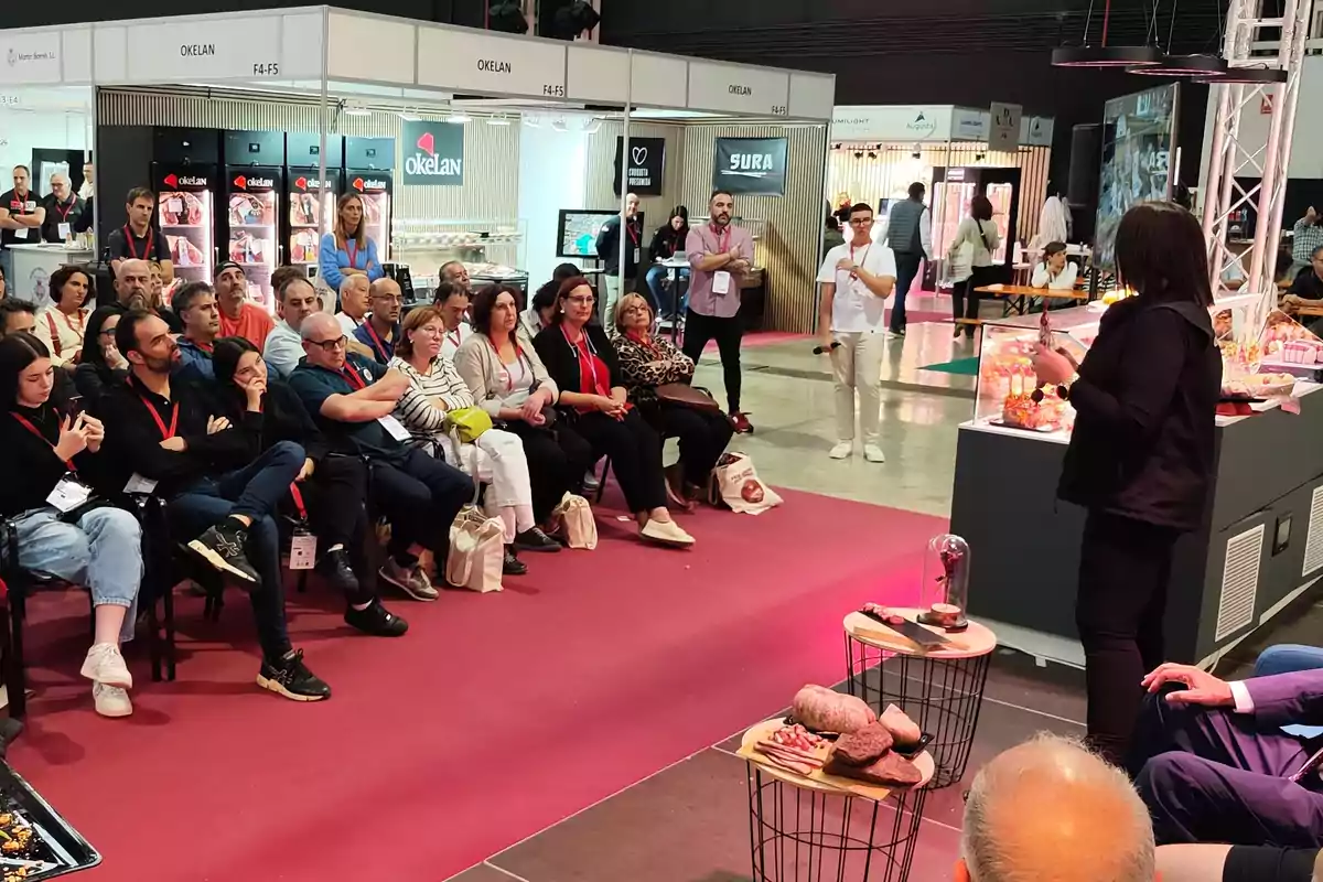 Un grupo de personas está sentado en sillas atendiendo a una presentación en un evento o feria comercial con stands de exhibición al fondo.