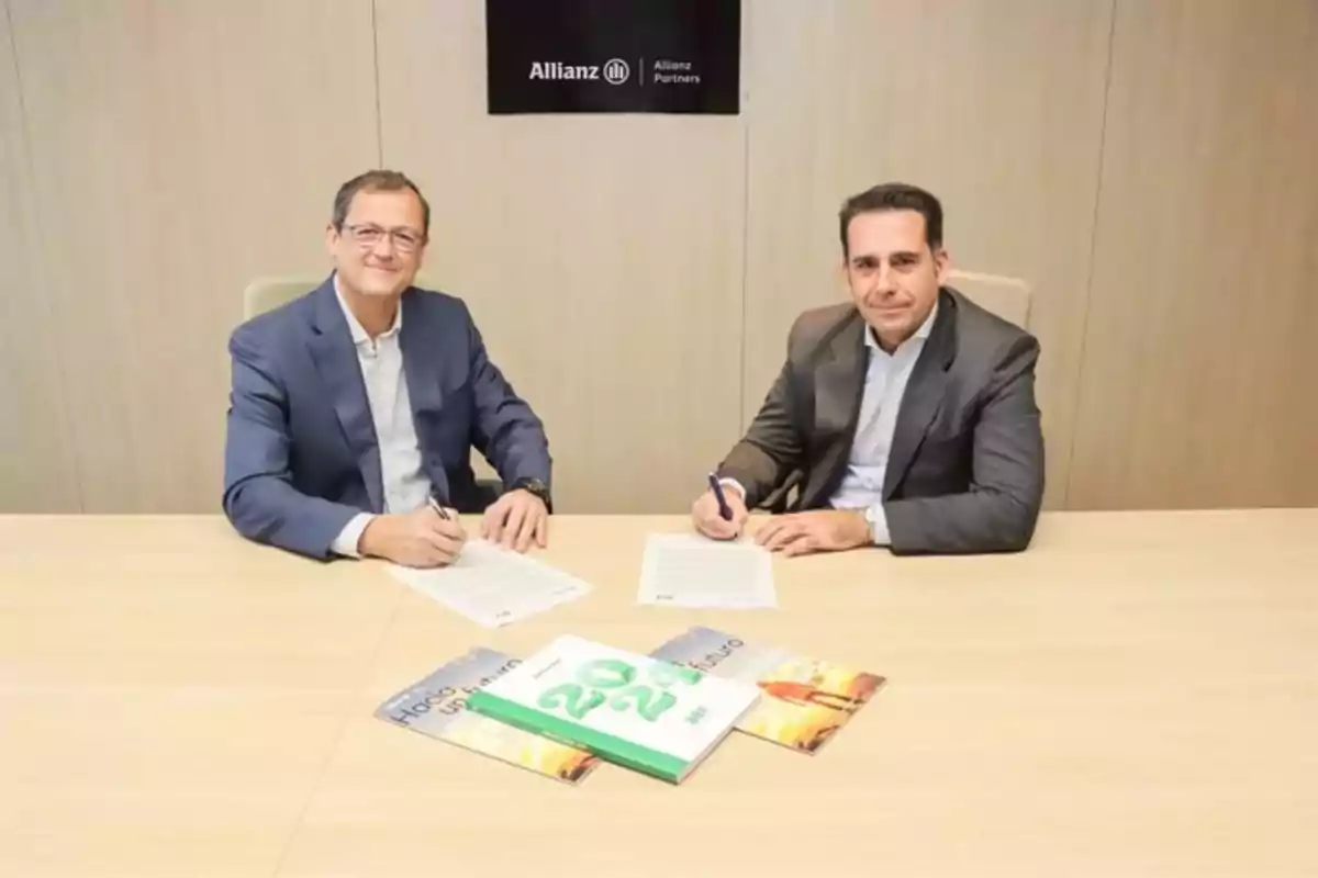 Dos hombres sentados en una mesa de oficina firmando documentos con una pared de fondo que tiene un logotipo de Allianz y varios folletos sobre la mesa.