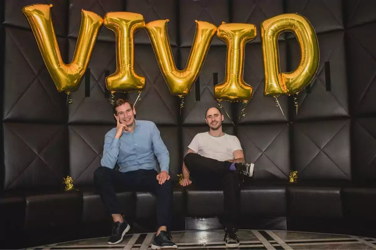 Dos personas sentadas en un sofá negro con globos dorados que forman la palabra "VIVID" sobre ellos.