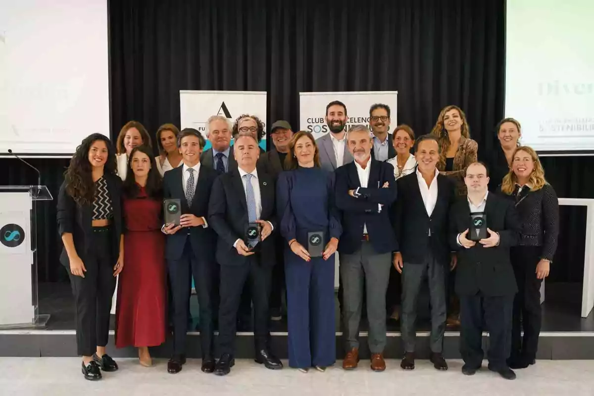 Un grupo de personas posando en un evento, algunas sosteniendo premios, con un fondo de pancartas y un podio.