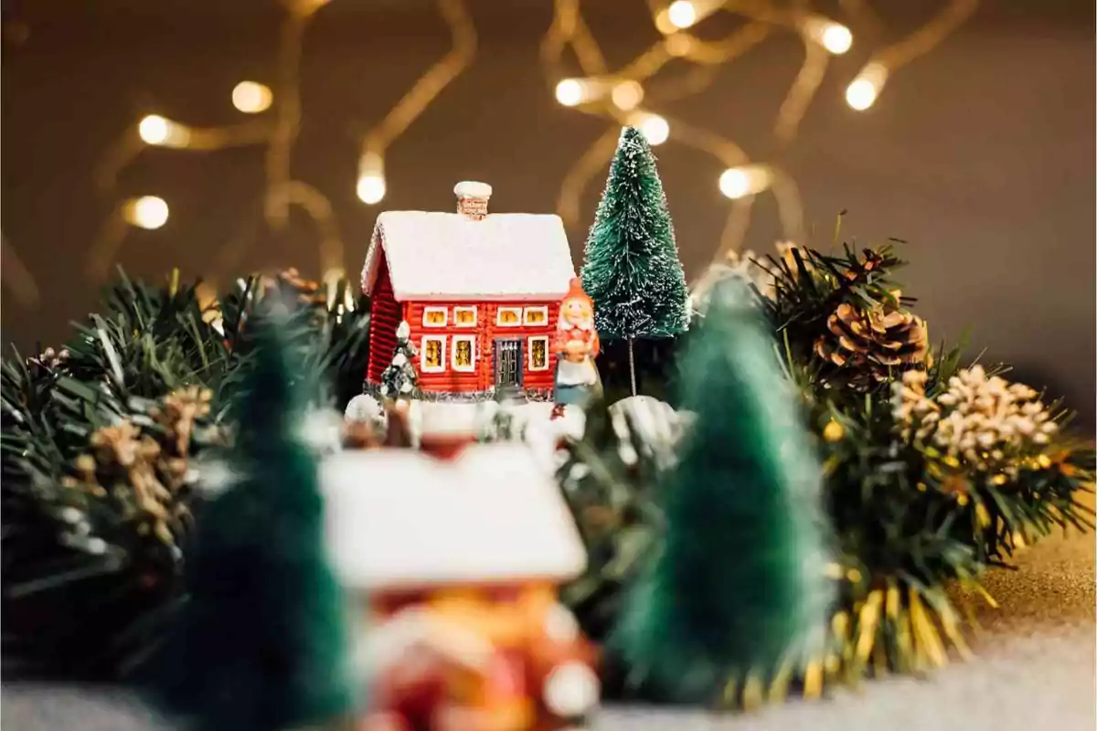 Una casa en miniatura con techo nevado rodeada de árboles y luces cálidas de fondo.