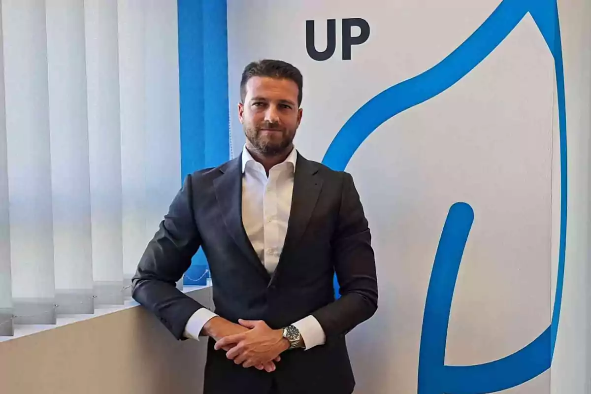 Un hombre con traje oscuro y camisa blanca está de pie frente a una pared con un diseño azul y la palabra "UP".