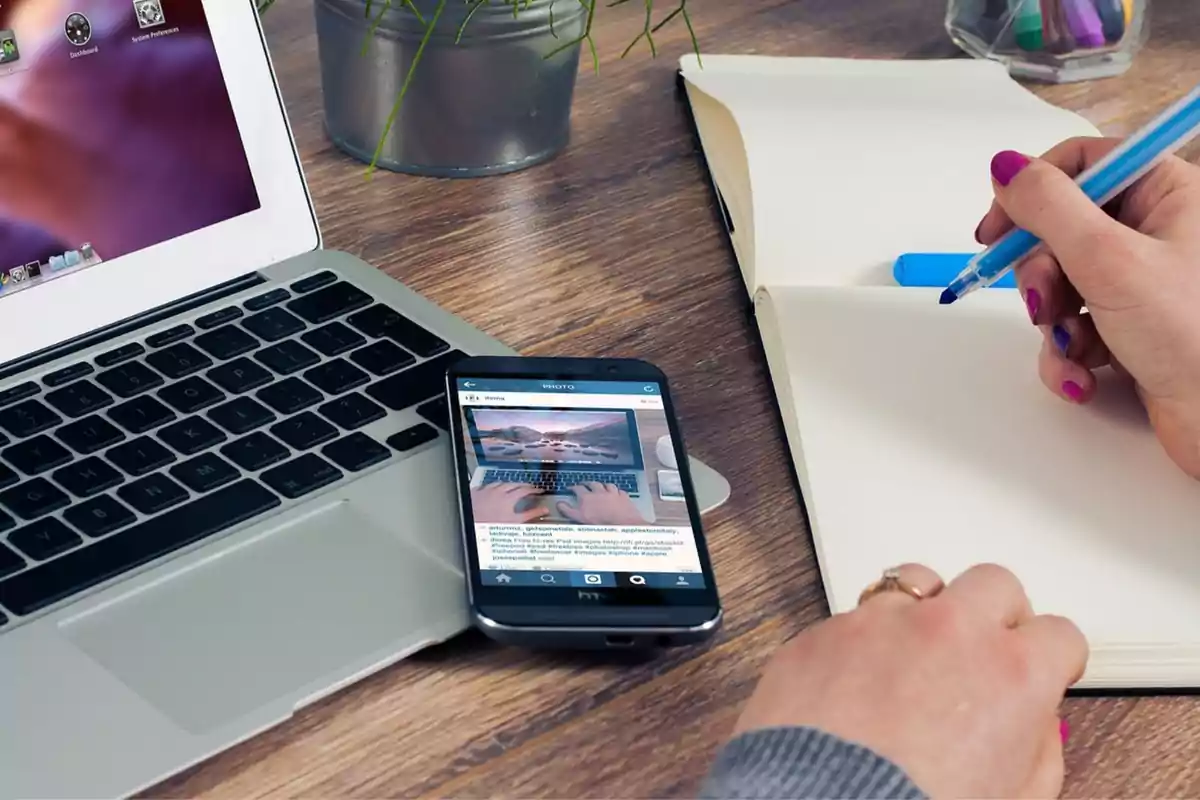 Persona escribiendo en un cuaderno junto a una computadora portátil y un teléfono móvil sobre un escritorio de madera.