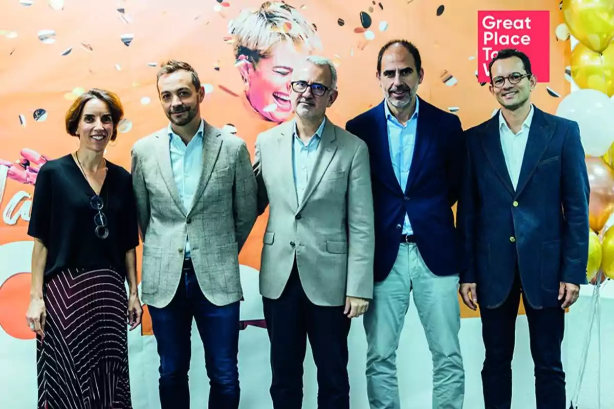 Cinco personas posan juntas frente a un fondo naranja con globos dorados y blancos y un cartel que dice "Great Place to Work".