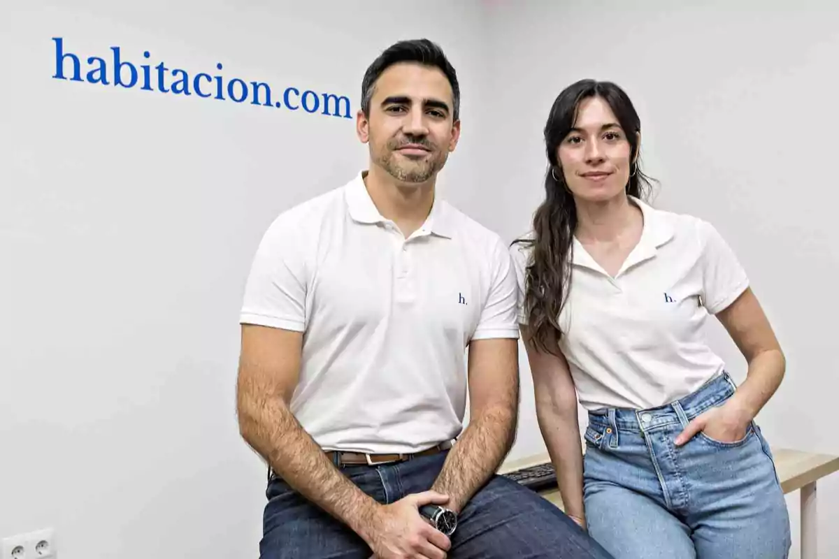 Dos personas con camisetas blancas posan frente a una pared blanca con el texto "habitacion.com" en azul.